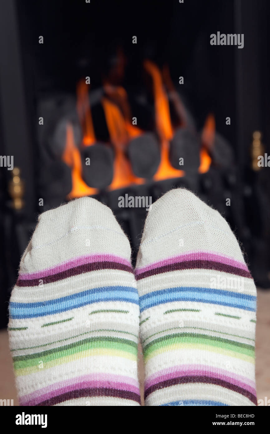 Les pieds d'une femme dans une paire de chaussettes en laine chaude stripy réchauffement climatique en face d'un feu de gaz de charbon confortable avec des charbons ardents à la maison en hiver. Angleterre Royaume-uni Grande-Bretagne Banque D'Images
