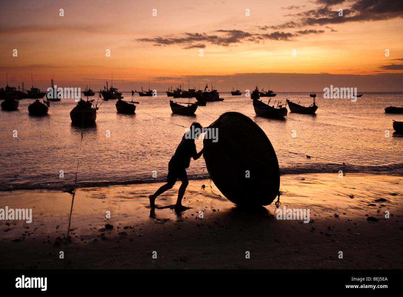 MUI NE, VIETNAM : Silhouette d'un homme roulant un bateau rond traditionnel vietnamien sur la plage au coucher du soleil. Mui Ne, Vietnam Banque D'Images