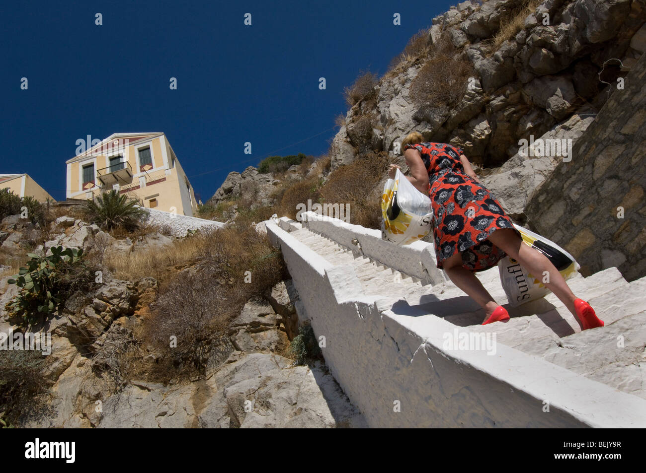 Une femme avec deux sacs de magasinage lourd raide grimpe étapes pour une maison sur l'île grecque de Symi Banque D'Images