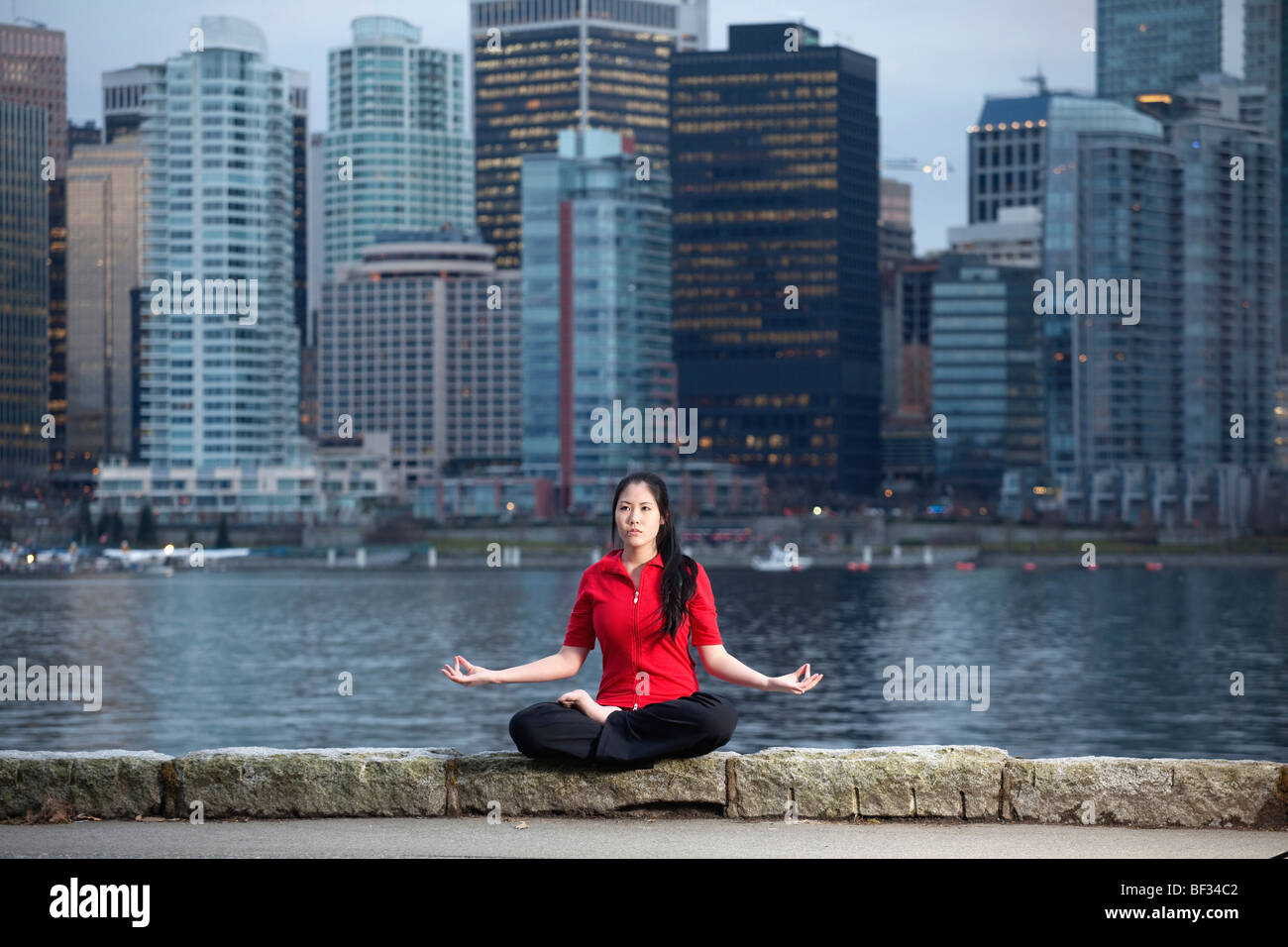 Femme Asiatique n'yoga Lotus Padmasana posent le long du sentier côtier du parc Stanley avec les toits de la ville au-delà, Vancouver, BC, Canada Banque D'Images