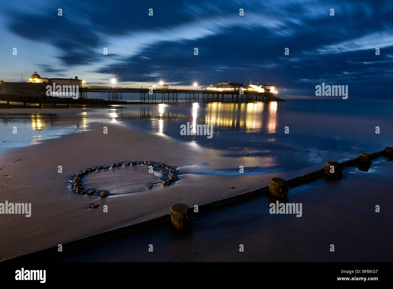 Une longue exposition de jetée de Cromer, au crépuscule avec des lumières se reflétant dans la mer et pebble circle dans le sable. Banque D'Images