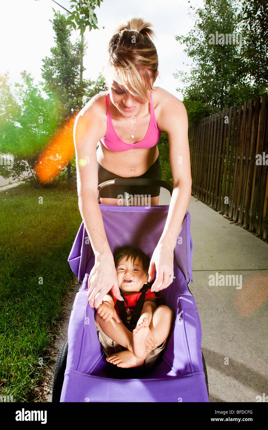 Une edgy-athlétique, à la jeune femme s'arrête de jouer avec un bébé dans sa poussette pendant le passage sur un trottoir de banlieue. Banque D'Images