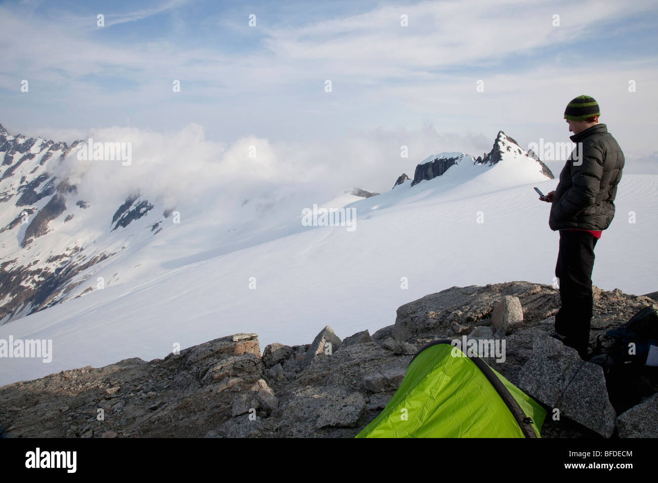 Jeune grimpeur vérifie son téléphone cellulaire pour la couverture tout en se tenant sur un affleurement rocheux sur un glacier avec des angles prononcés dans la distance Banque D'Images