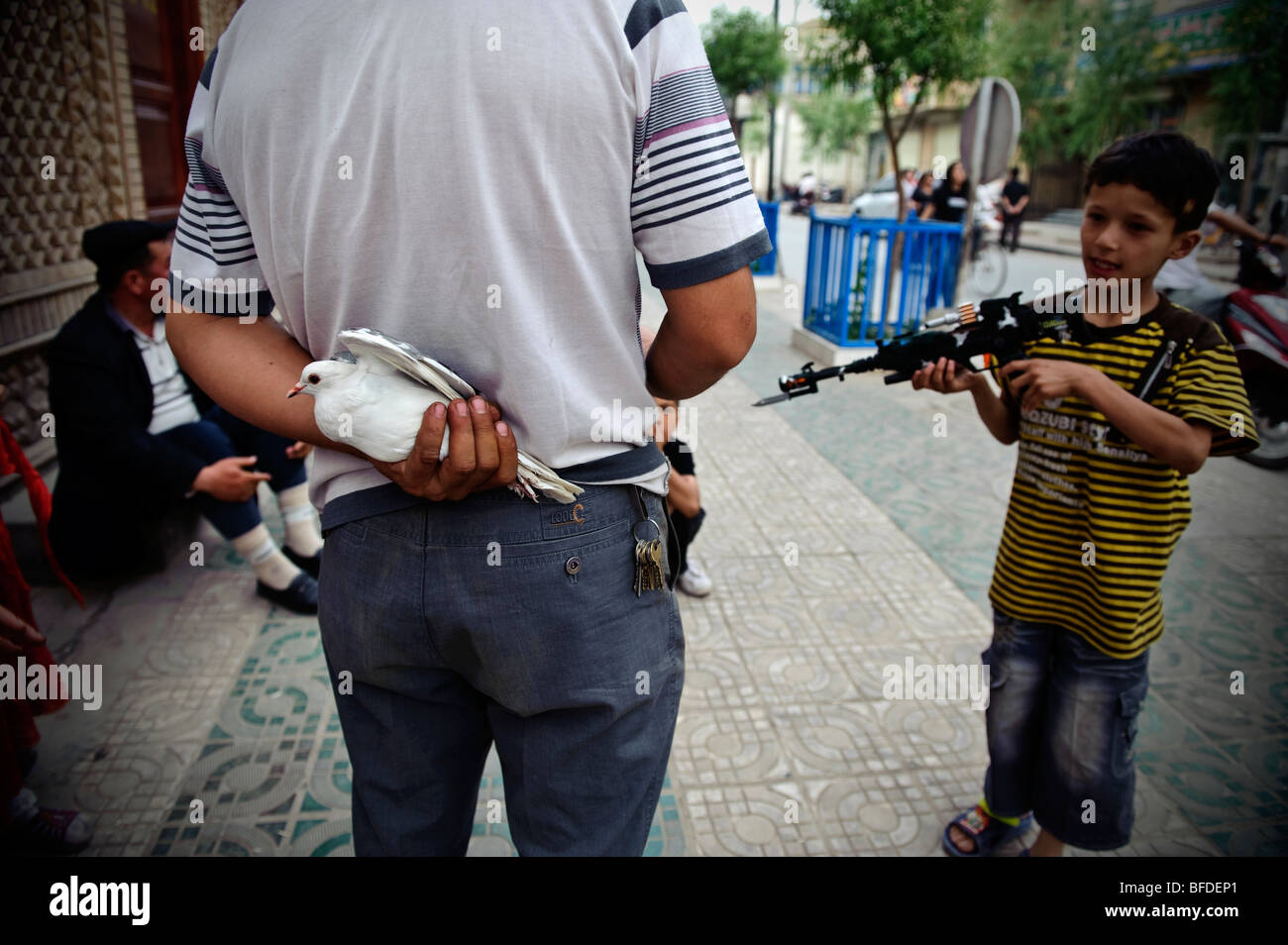 Un homme Ouïghour de la protection d'un pigeon blanc à partir d'un groupe de garçons jouant avec des armes en plastique sur une rue de Kashgar, Xinjiang, Chin Banque D'Images