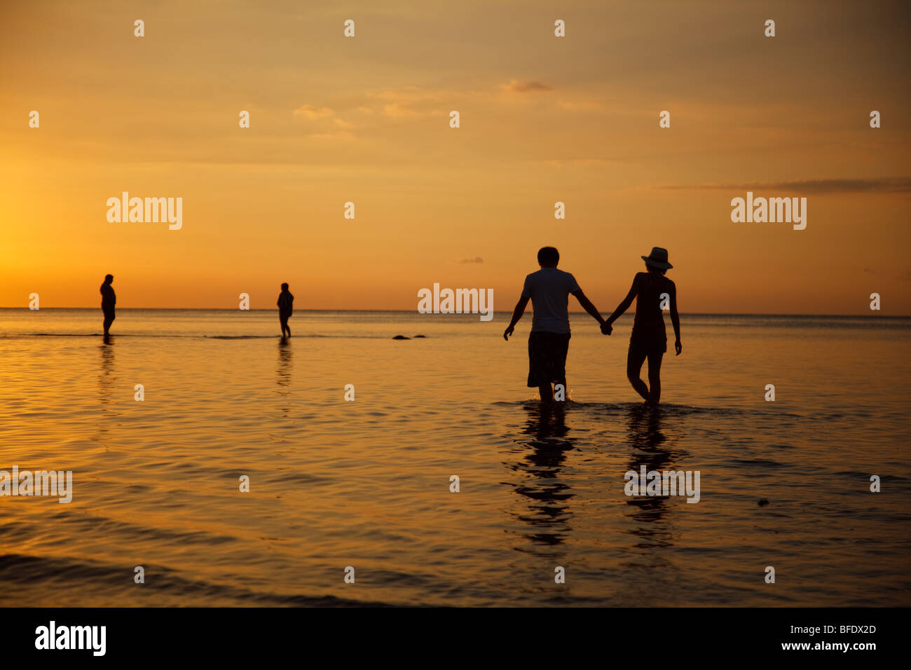 Un jeune couple de vacanciers détient les mains et entre dans la mer lors d'un magnifique coucher de soleil sur la plage de Lovina, Bali Banque D'Images