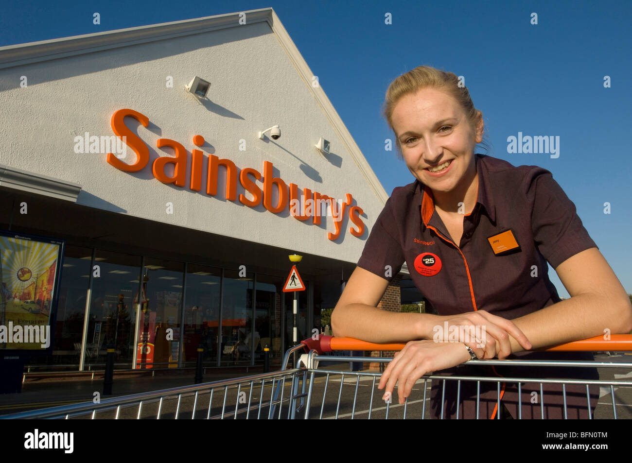 Supermarché Sainsbury's . Smiling girl en uniforme avec chariot à l'extérieur d'un magasin. Banque D'Images