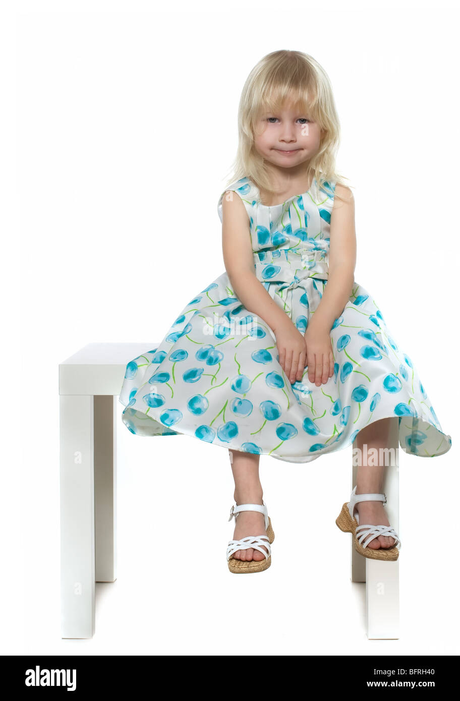 Petite fille blonde en robe bleu-blanc est assis sur chaise blanche, isolated on white Banque D'Images