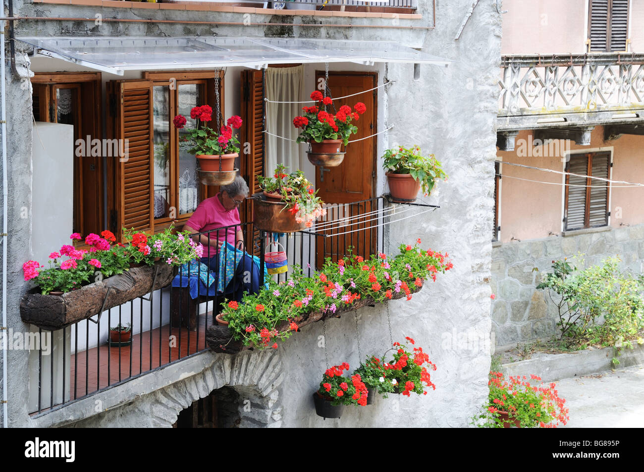 Vieille dame la couture sur balcon entouré par les jardinières pots Fleurs rouge rose géranium Pont St Martin Vallée d'Aoste Italie Banque D'Images
