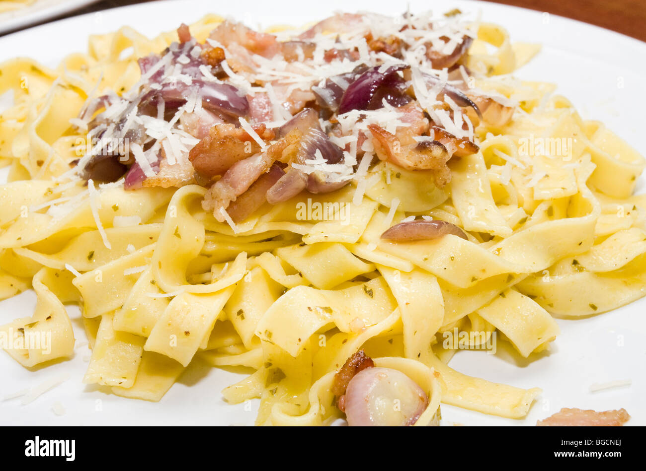 Assiette de tagliatelles sautées dans l'huile d'olive et au pesto avec lard grillé et oignon rouge. Garni de fromage parmesan râpé. Banque D'Images