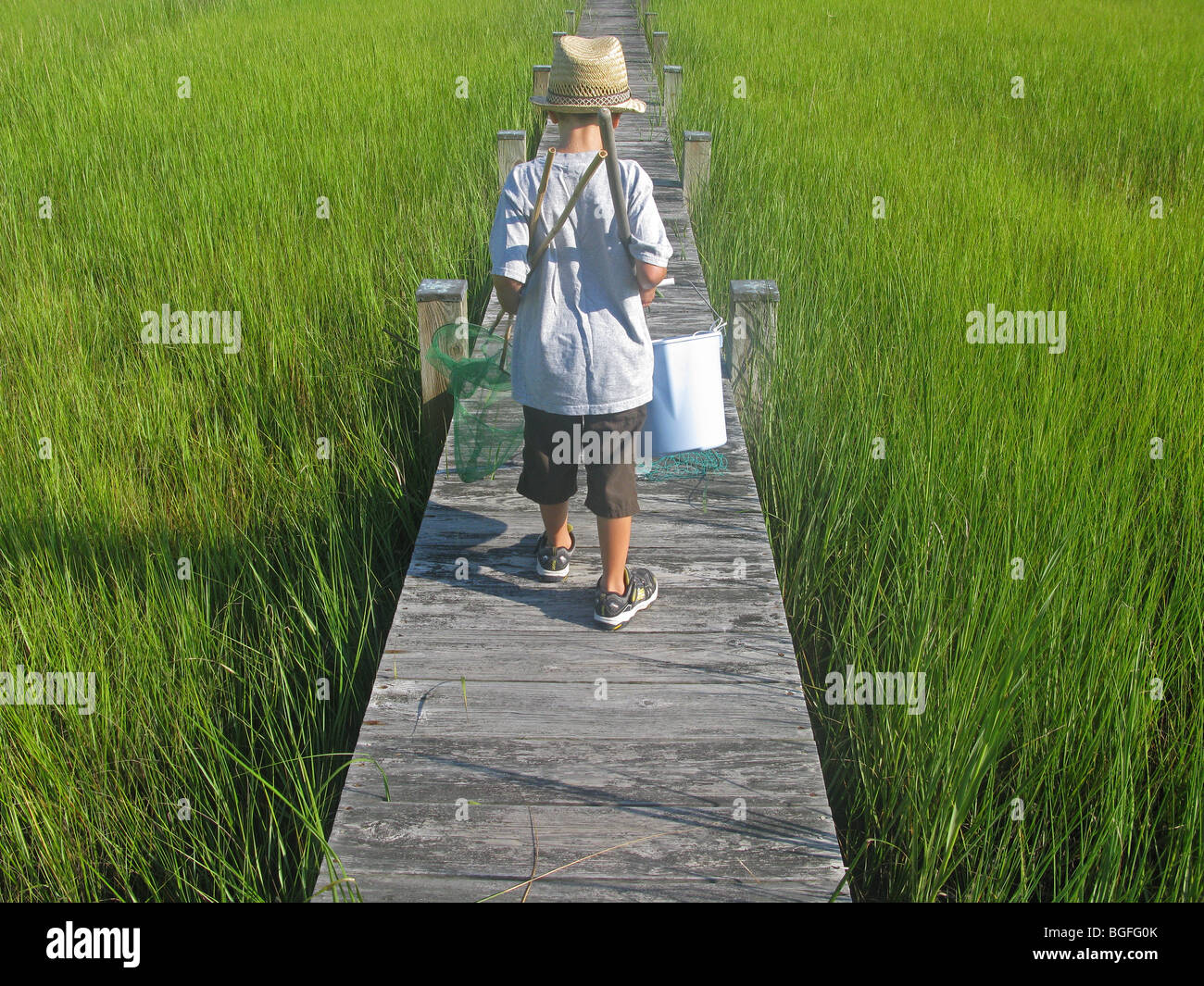 Un jeune garçon marchant le long d'une jetée en bois avec canne à pêche, filet et un seau. Banque D'Images