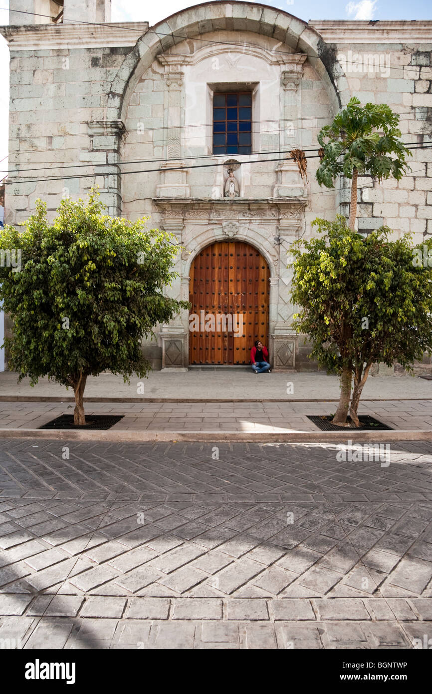 Sloppy teenage girl sitting in porte de la vieille église en pierre bordée par un trottoir à feuilles vertes ficus dans la ville d'Oaxaca au Mexique Banque D'Images