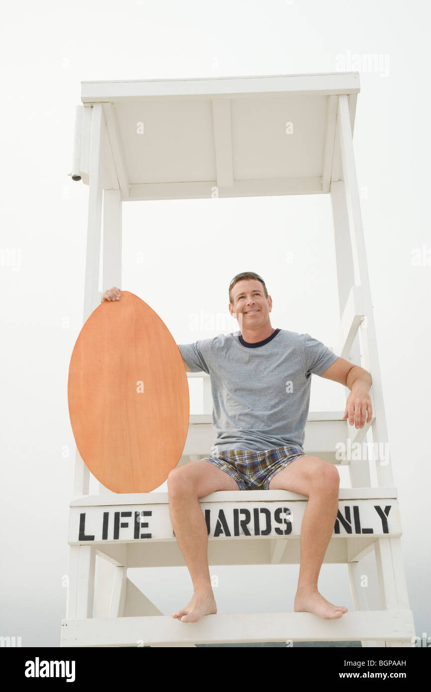 Homme mûr assis sur un lifeguard hut et tenant une planche de surf Banque D'Images