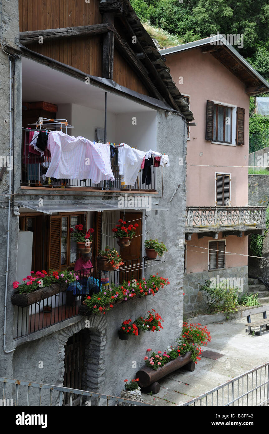Vieille dame la couture sur balcon entouré par les jardinières pots de fleurs rose rouge géraniums dans vieille maison Pont St Martin Aosta Banque D'Images