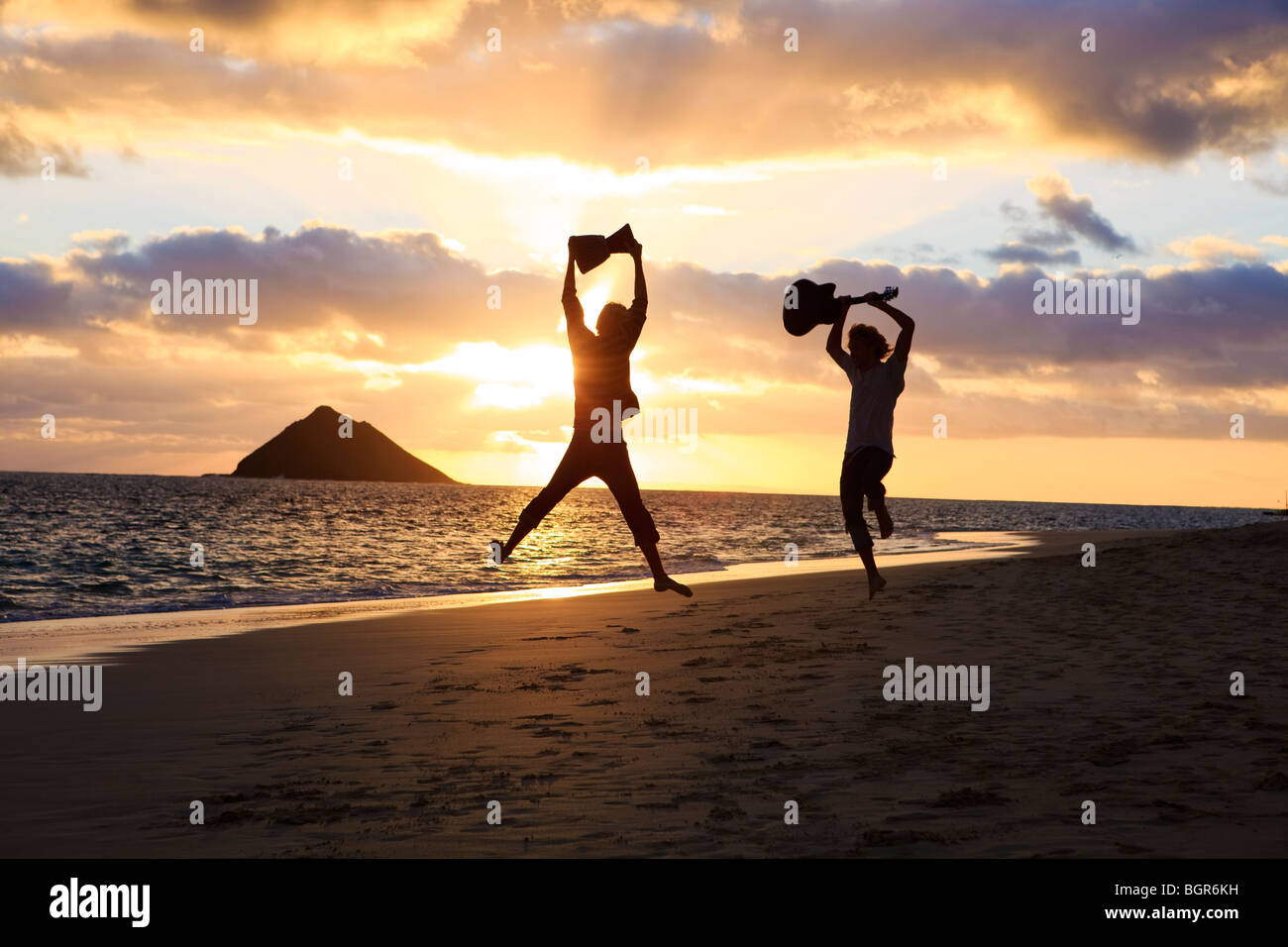 Silhouette de frères jumeaux avec batterie et guitare en sautant en l'air au lever du soleil sur une plage de Hawaï Banque D'Images