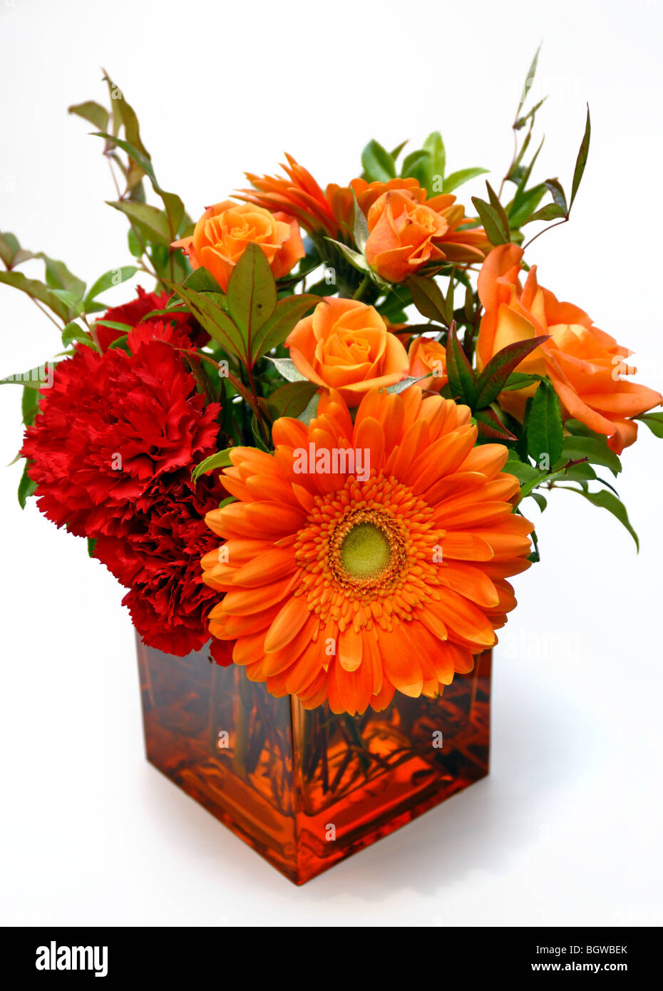 Un arrangement de fleurs orange colorés contenant des marguerites et roses, dans un vase en verre carré. Banque D'Images