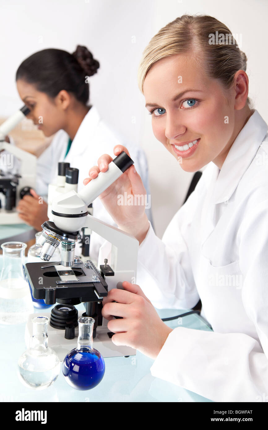 Une femme blonde chercheur scientifique ou médical ou un médecin à l'aide de son microscope dans un laboratoire avec son collègue asiatique Banque D'Images