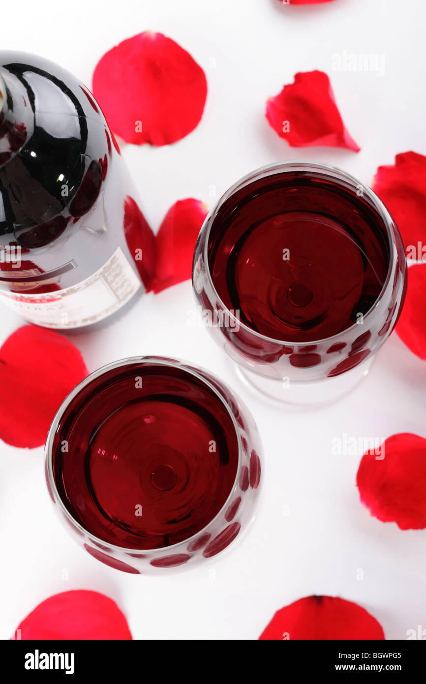 Une paire de verres de vin remplis de vin rouge entourée de pétales de rose rouge sur un fond blanc et une bouteille de vin Banque D'Images
