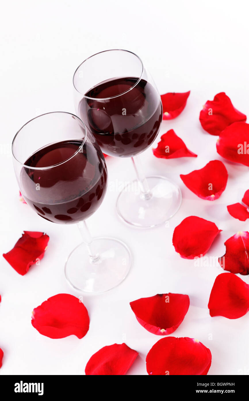 Une paire de verres de vin remplis de vin rouge entourée de pétales de rose rouge sur un fond blanc Banque D'Images
