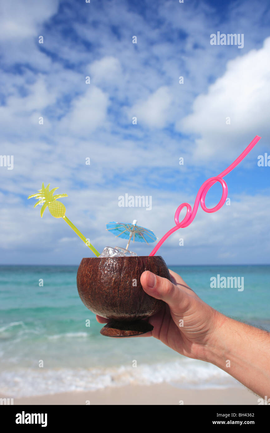 La main d'un homme tenant un cocktail tropical dans une coque de noix de coco sur une plage tropicale déserte Banque D'Images