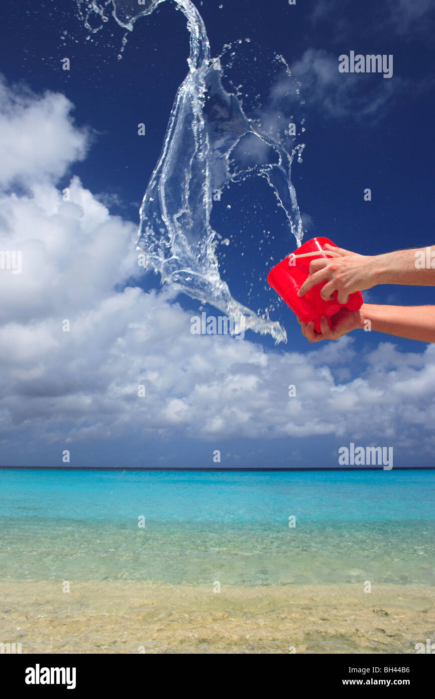Les mains d'un homme jetant un jouet rouge seau plein d'eau dans l'air sur une plage tropicale déserte Banque D'Images