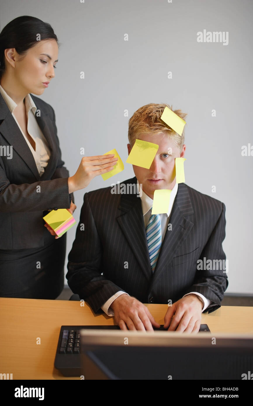 Une femme d'en plaçant les notes de papier sur le visage pour un homme d'affaires, comme il est assis à un bureau à travailler sur un ordinateur dans un bureau Banque D'Images
