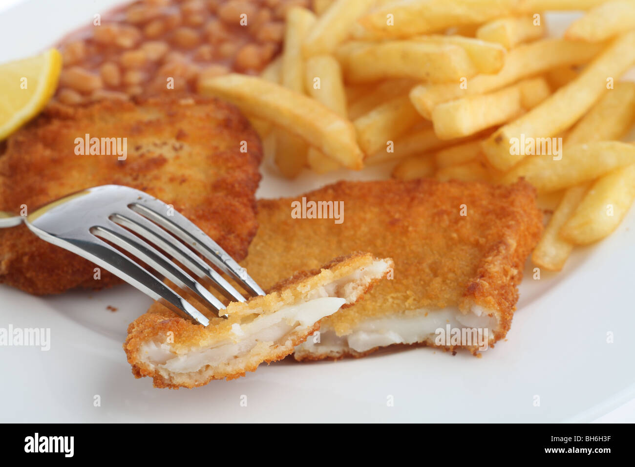 Un morceau de filet de poisson pané sur une fourchette, avec le repas de haricots cuits, chips (frites) et un quartier de citron derrière. Banque D'Images