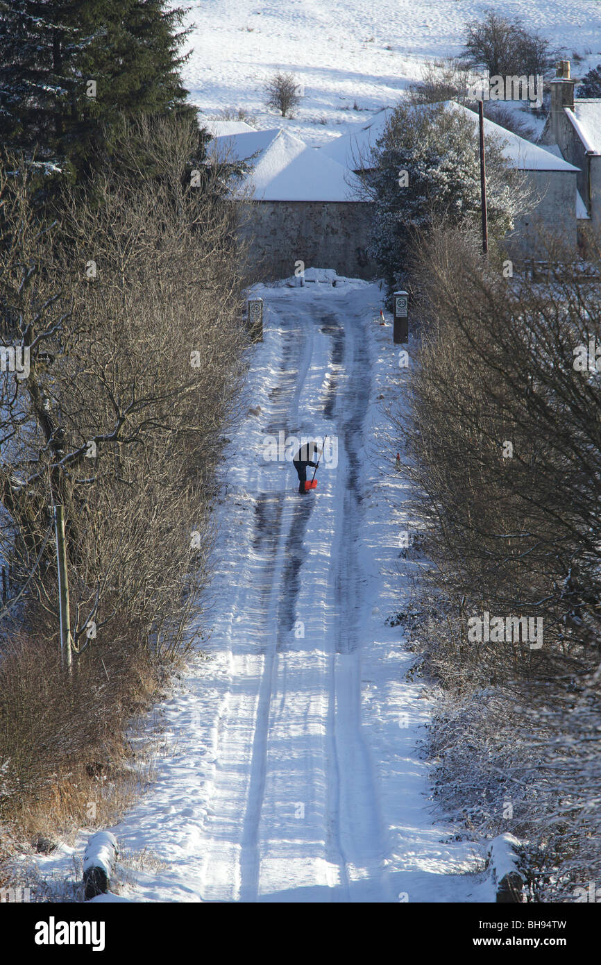 Homme défrichant la neige avec une pelle d'une longue route vallonnée en campagne en hiver, Écosse, Royaume-Uni Banque D'Images