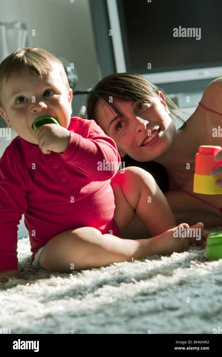 Jeune femme et bébé garçon assis sur un tapis à mâcher sur et jouer avec des jouets Banque D'Images