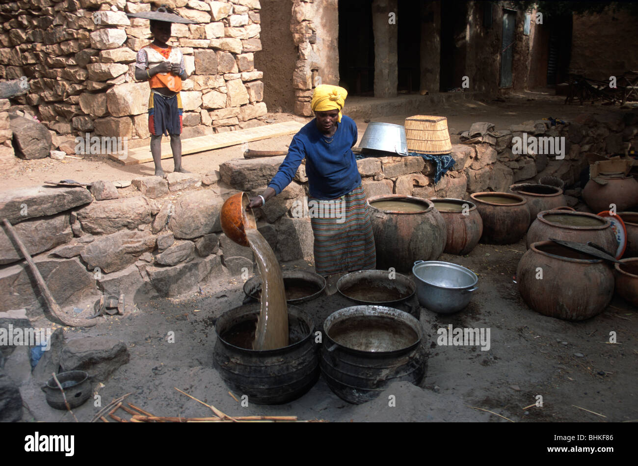 Un Malien woman pouring bière de mil brassée en partie dans un pot, dans Tereli, Mali, alors qu'un garçon montres avec un couvercle sur la tête. Banque D'Images