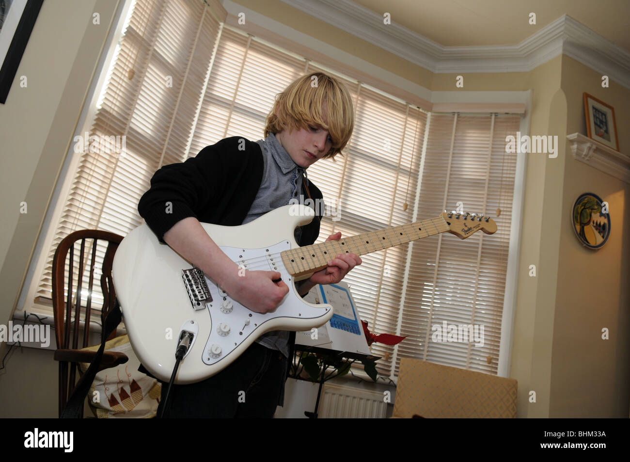 Teenage boy pratiquer le blues dans son salon sur une guitare Fender Stratocaster crème encadrée par une baie vitrée en toile Banque D'Images