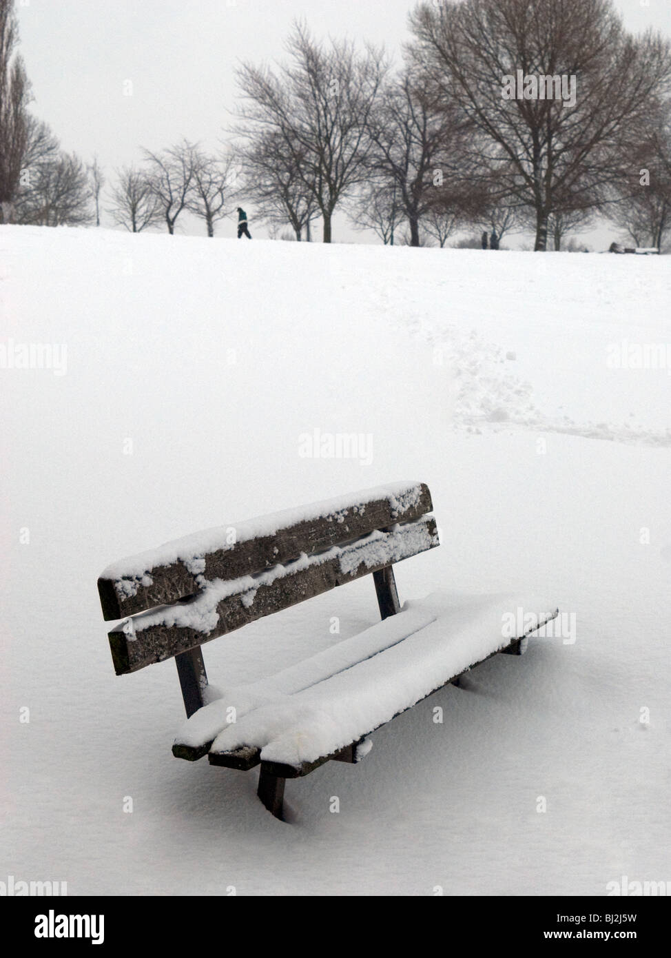 Banc en bois en dévers Park, South East London, après de fortes chutes de neige avec des arbres et des piétons en arrière-plan Banque D'Images