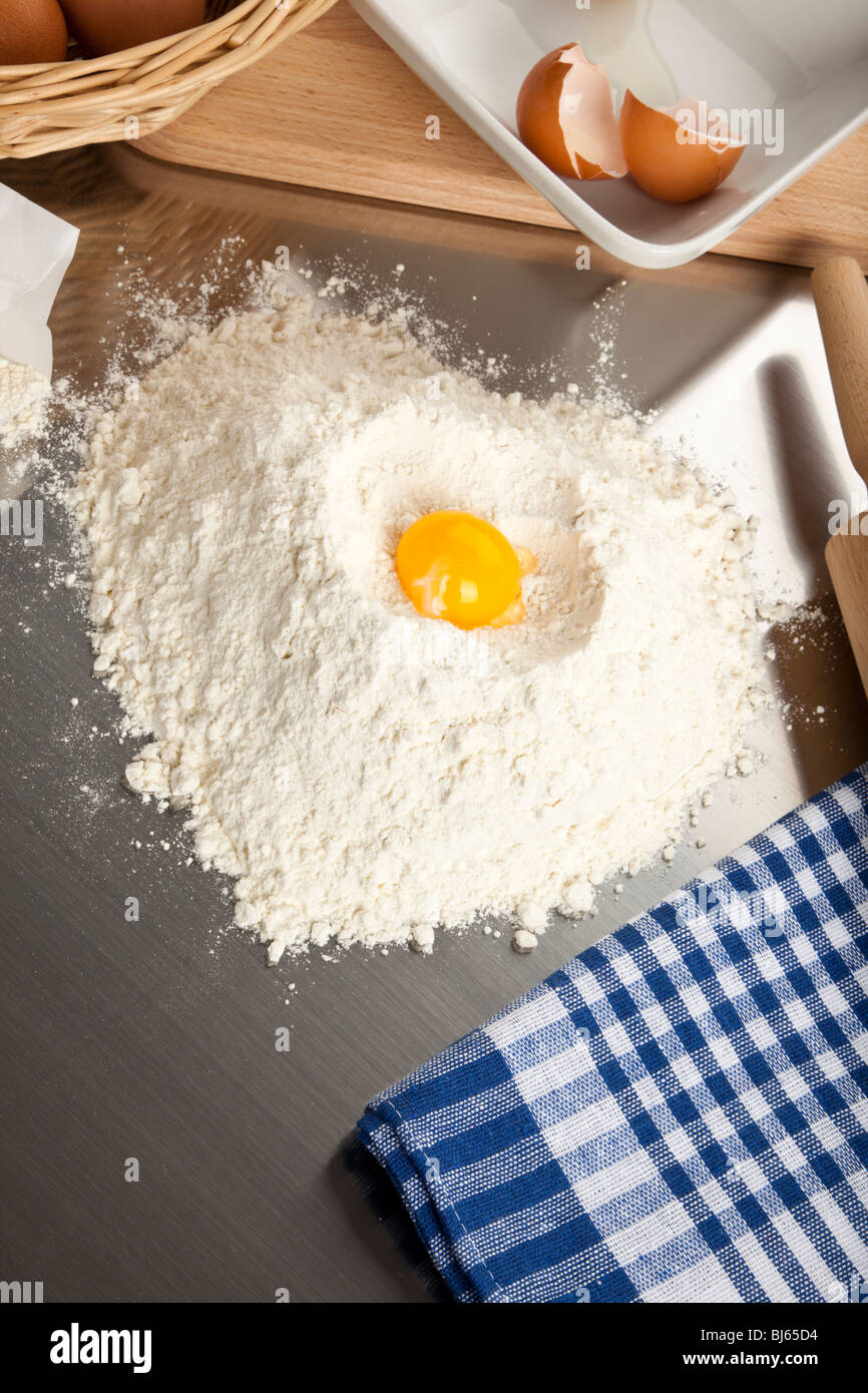 D'œuf sur le tas en forme de coeur de la farine de blé sur la surface en métal brossé, avec une serviette de cuisine, rouleau à pâtisserie, des œufs et de la coquille brisée Banque D'Images