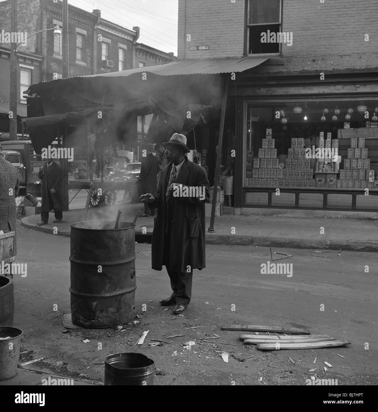 Bien habillé d'un homme afro-américain se réchauffe sa main sur un baril fire à Philadelphie au début des années 1950. pauvres devanture Banque D'Images