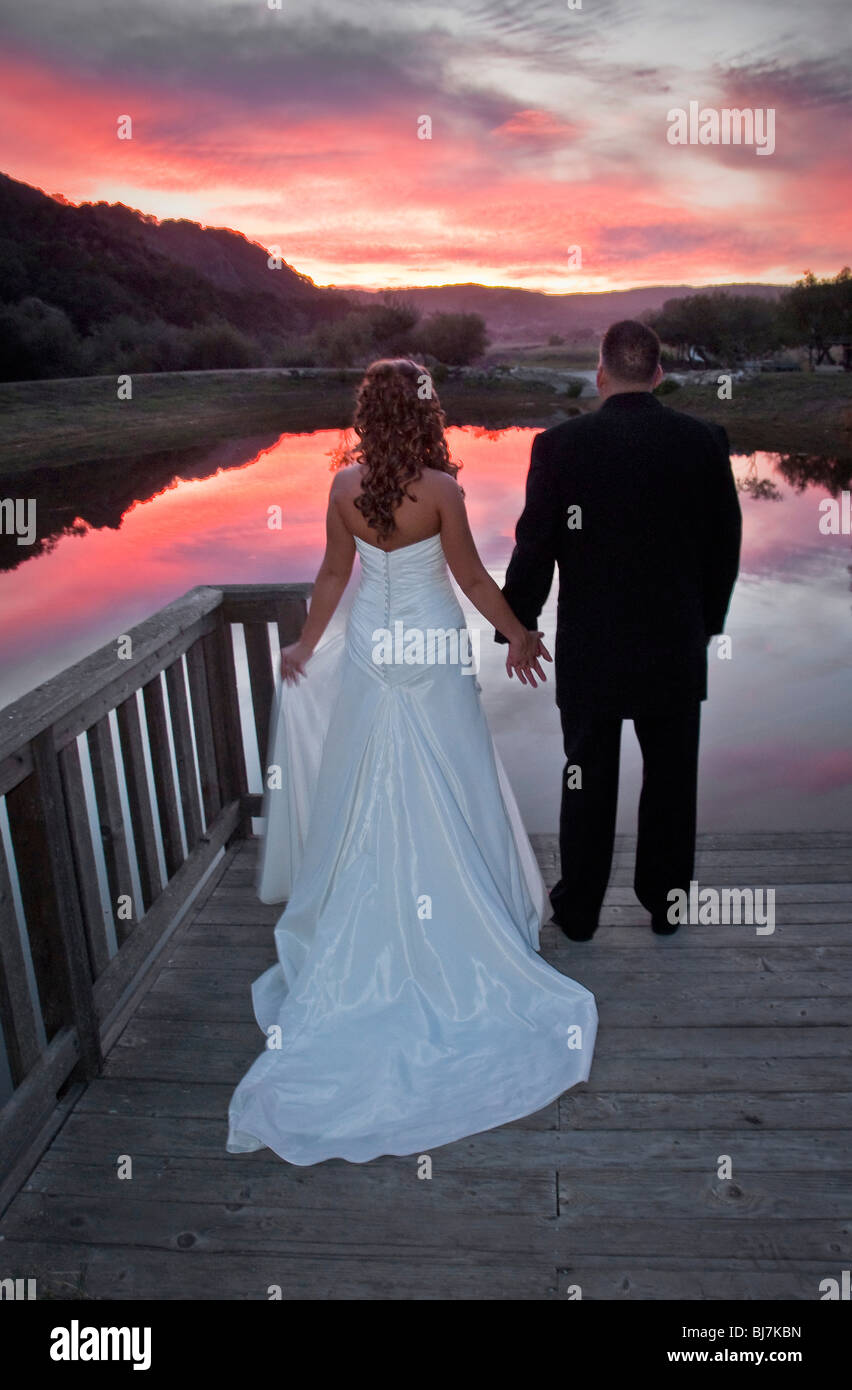 Bride and Groom tenir la main et regarder un lac au coucher du soleil. Banque D'Images