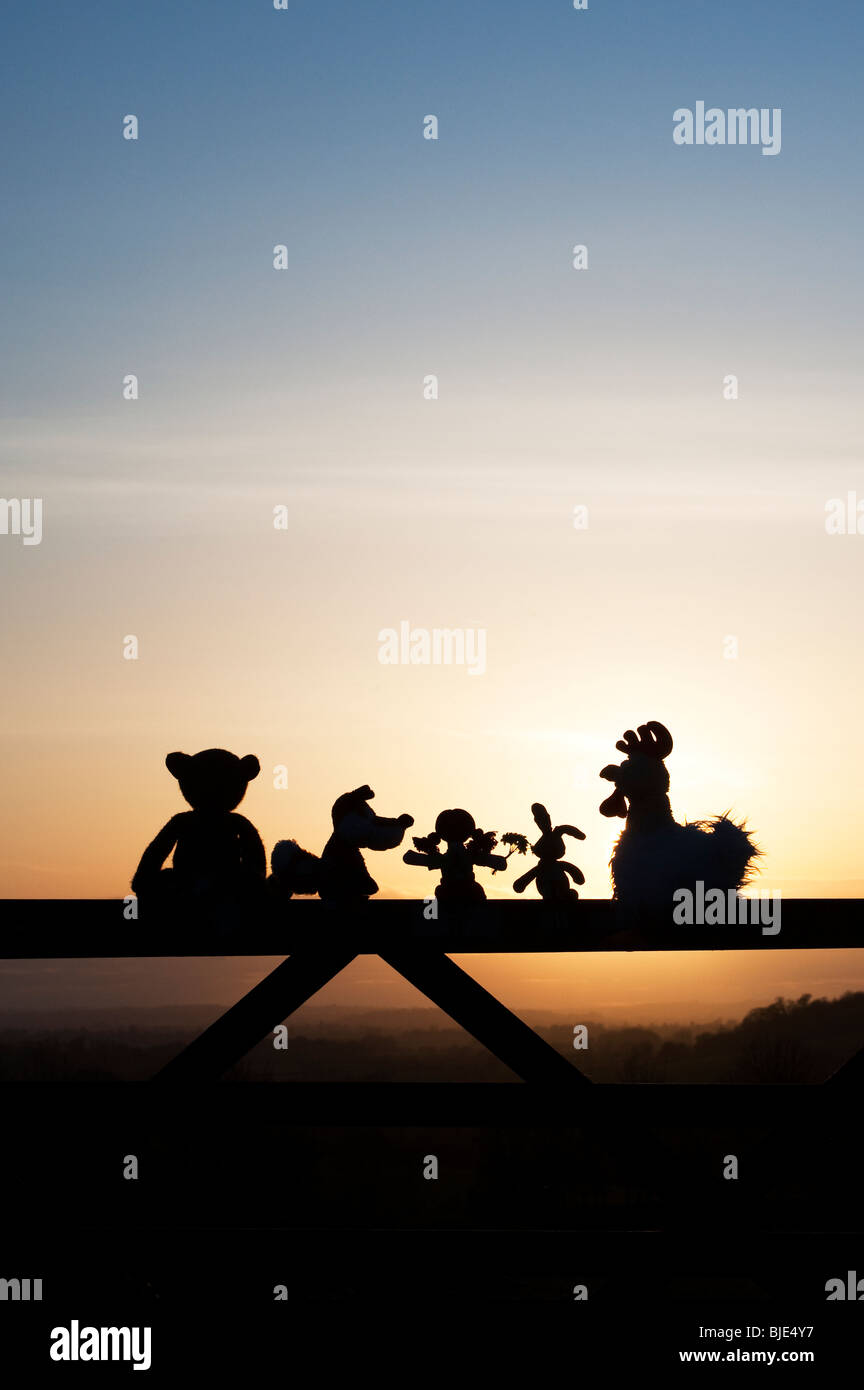 Poupée de chiffon, poulet, lapin, renard et l'ours peluches assis sur une barrière au coucher du soleil dans la campagne anglaise. Silhouette Banque D'Images