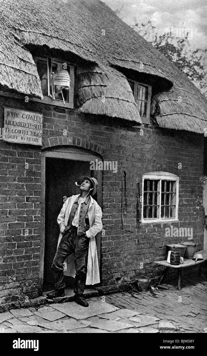 Un homme fumant une pipe à l'extérieur d'une boutique, Worcestershire, c1922.Artist : AW Cutler Banque D'Images