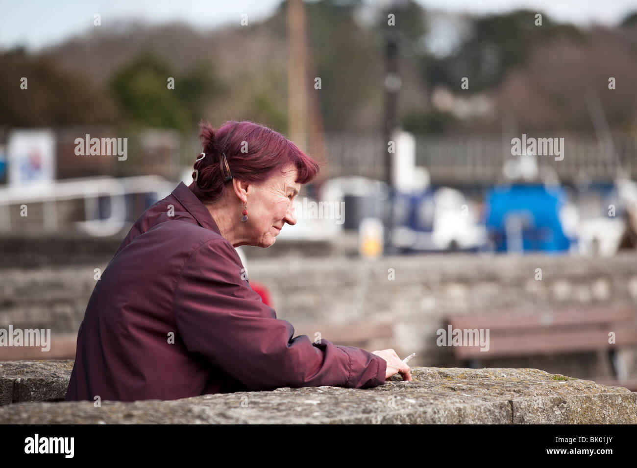 Une vieille dame au manteau marron avec les cheveux marron appuyé contre un mur bas fumer une cigarette Banque D'Images