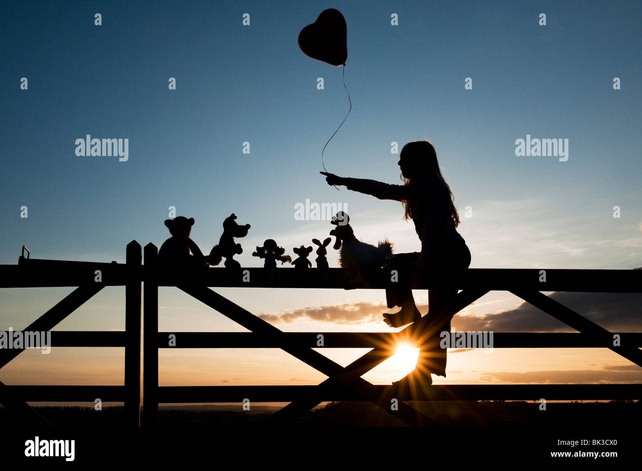 Girl holding a balloon avec une poupée de chiffon, poulet, lapin, renard et l'ours peluches assis sur une barrière au coucher du soleil. Silhouette Banque D'Images
