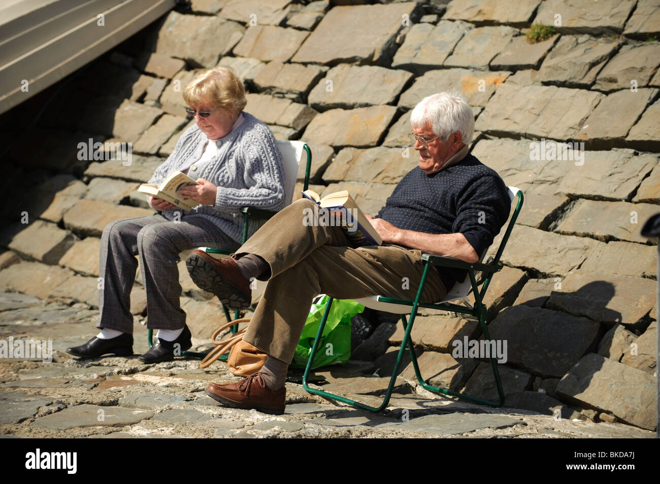 Un couple de retraités âgés assis au soleil la lecture de livres à New Quay, Ceredigion, pays de Galles, Royaume-Uni Banque D'Images