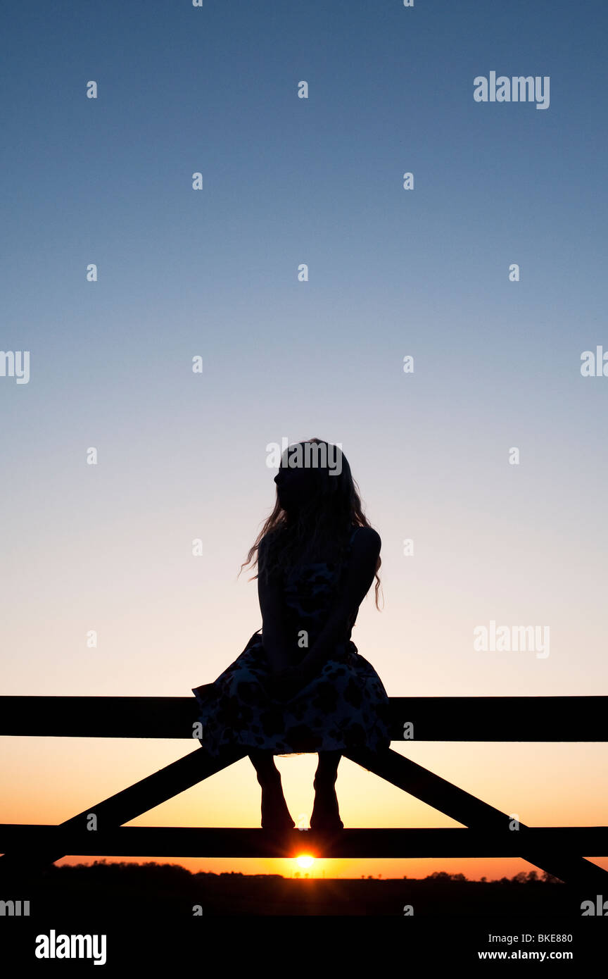 Jeune fille assise sur une porte au coucher du soleil. Silhouette Banque D'Images