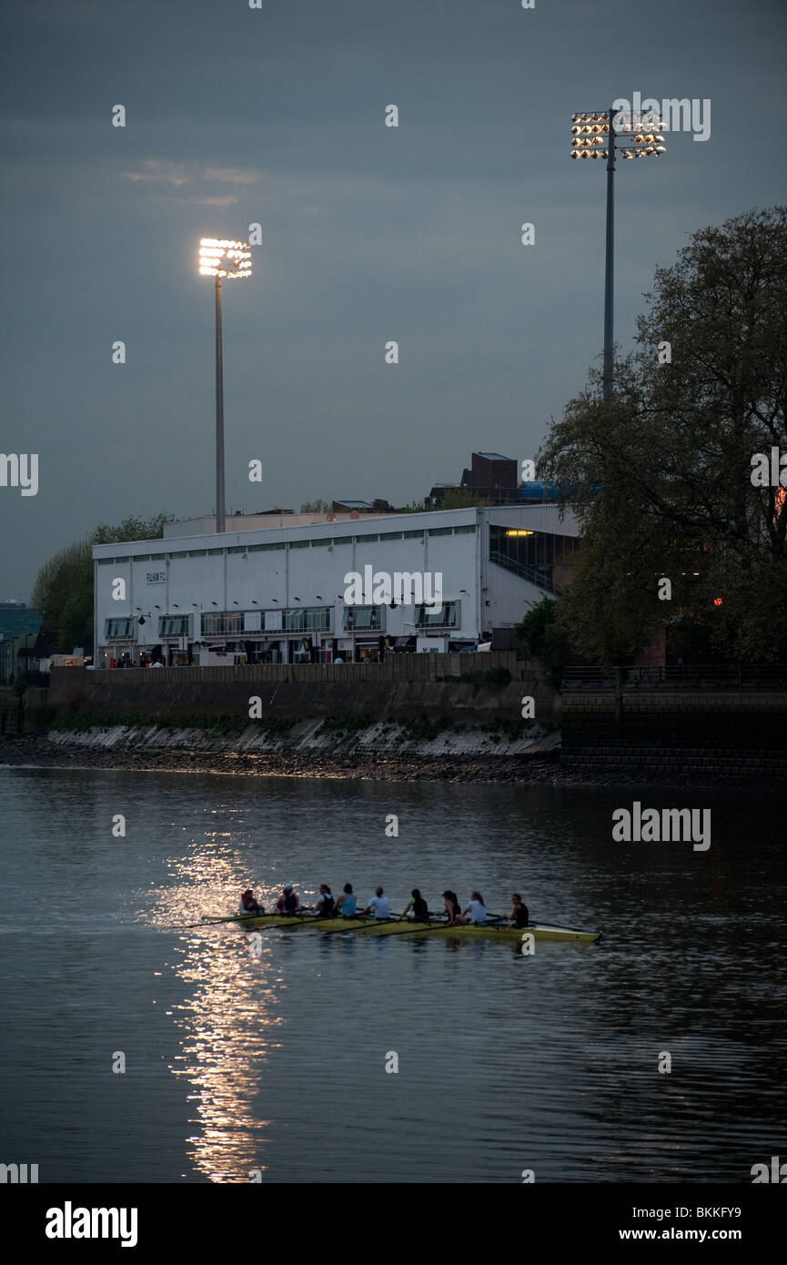 Un bateau plein de rameurs glisse passé Craven Cottage terrain de football au crépuscule que la lueur des projecteurs sur la rivière Thames, London. Banque D'Images