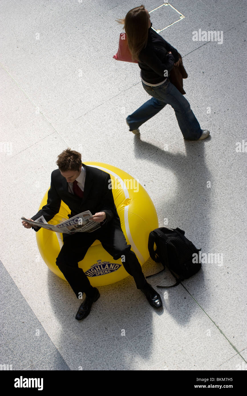 Un voyageur se détendant sur une chaise gonflable, dans le cadre de la promotion Highland Spring boissons gazeuses tout en lisant le journal Metro gratuit. Liverpool Street Londres Banque D'Images