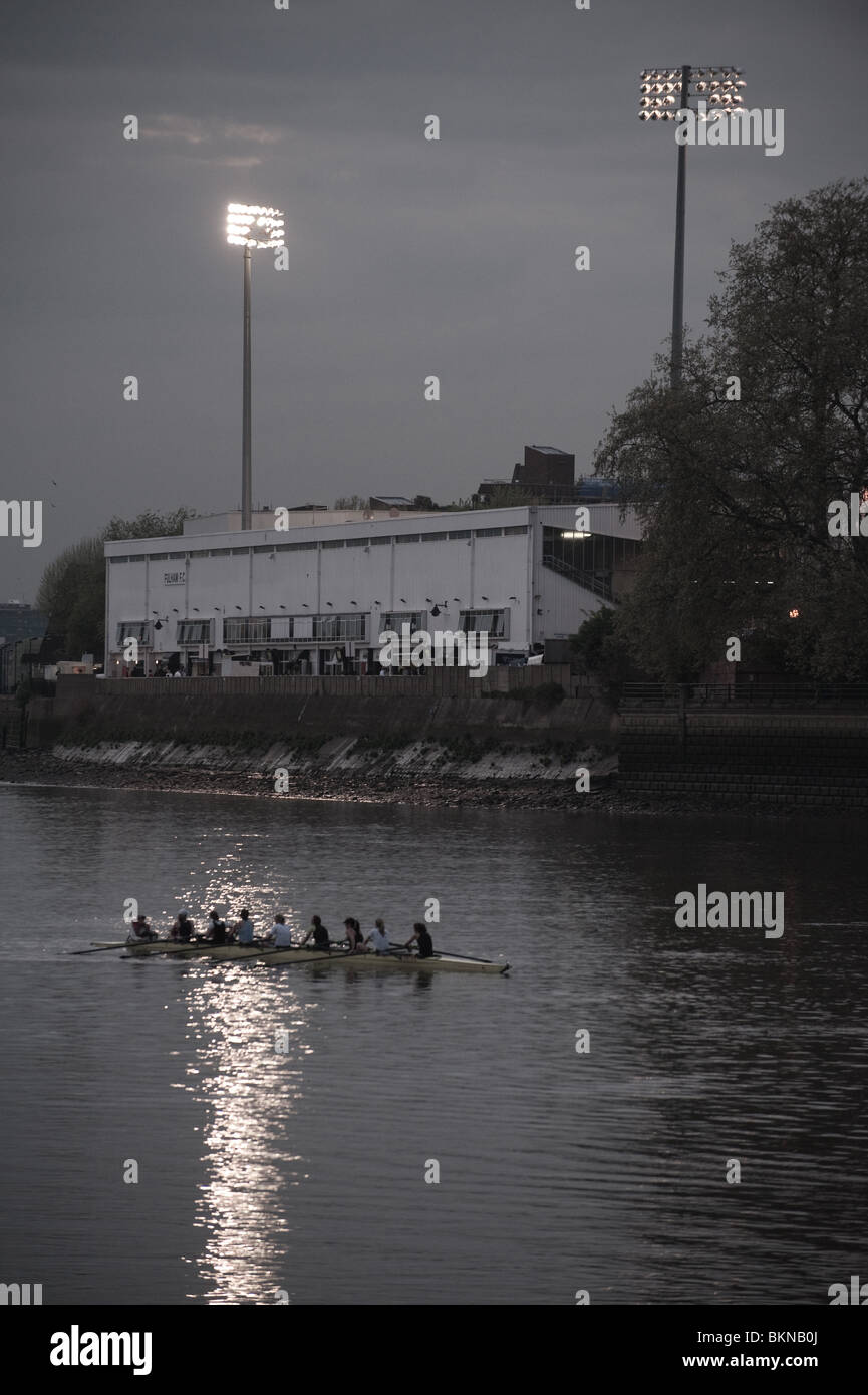 Un bateau plein de rameurs glisse passé Craven Cottage terrain de football au crépuscule que la lueur des projecteurs sur la rivière Thames, London. Banque D'Images