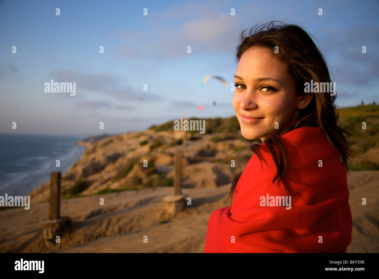 Femme dans la jeune vingtaine, enveloppé dans une couverture rouge sourit à la caméra pendant qu'elle regarde le parapente au coucher du soleil sur l'ocea Banque D'Images Femme dans la jeune vingtaine, enveloppé dans une couverture rouge sourit à la caméra pendant qu'elle regarde le parapente au coucher du soleil sur l'ocea Banque D'Images