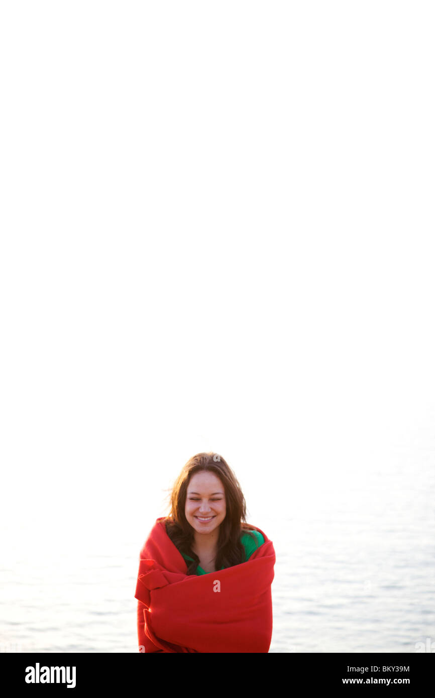Femme dans la jeune vingtaine, enveloppé dans une couverture rouge sourit avec ses yeux fermés sur la plage. Banque D'Images Femme dans la jeune vingtaine, enveloppé dans une couverture rouge sourit avec ses yeux fermés sur la plage. Banque D'Images