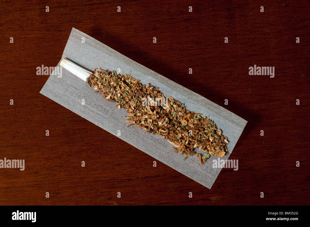 Un joint de cannabis et de mauvaises herbes (tabac) sur un papier à rouler sur une table en bois. Banque D'Images