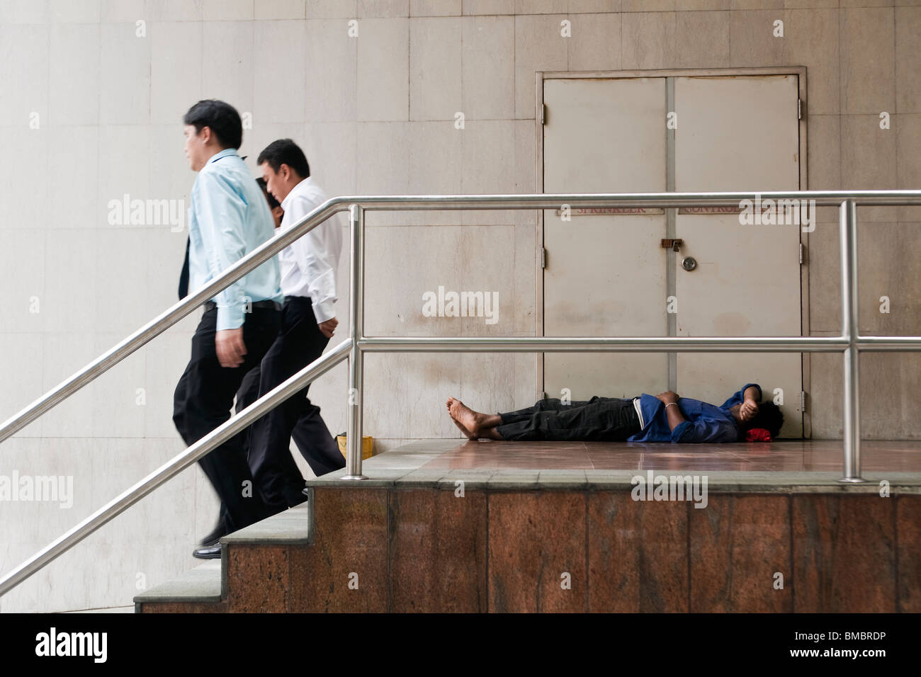 Les employés de bureau passent devant un travailleur migrant prendre une sieste au cours de sa pause déjeuner dans le quartier central des affaires à Singapour Banque D'Images