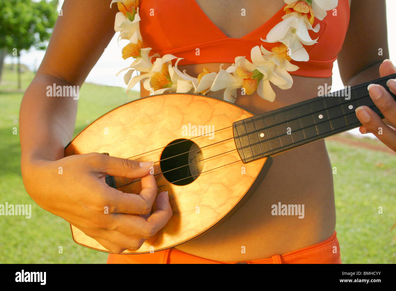Gros plan du hawaiian woman in bikini with orange orchid lei jouer ukulele ananas en parc. Honolulu, Oahu, HI Banque D'Images
