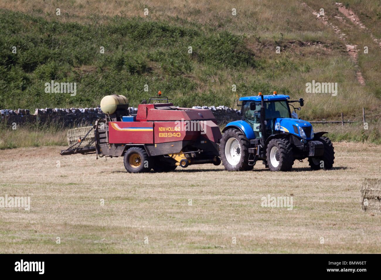 Tracteur agricole et du foin bleu collecte de la ramasseuse-presse sur les andains de foin, de l'agriculture dans la région ensoleillée du pays de Galles au Royaume-Uni. Vue côté horizontal. Banque D'Images