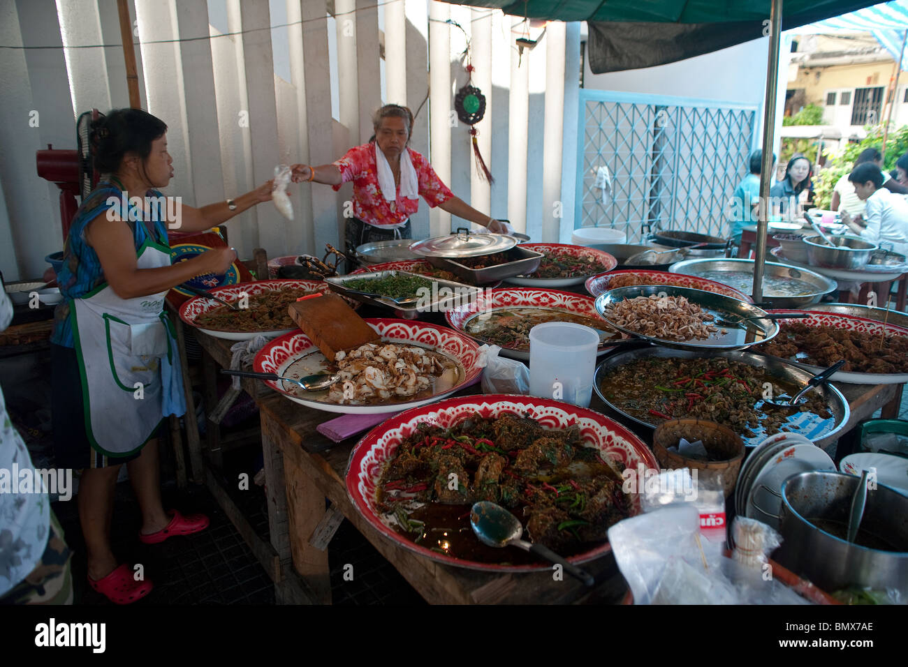 Food avec diverses spécialités et plaques de cuisine thaï et deux femmes en service alors que les clients de manger sont visibles à droite Banque D'Images